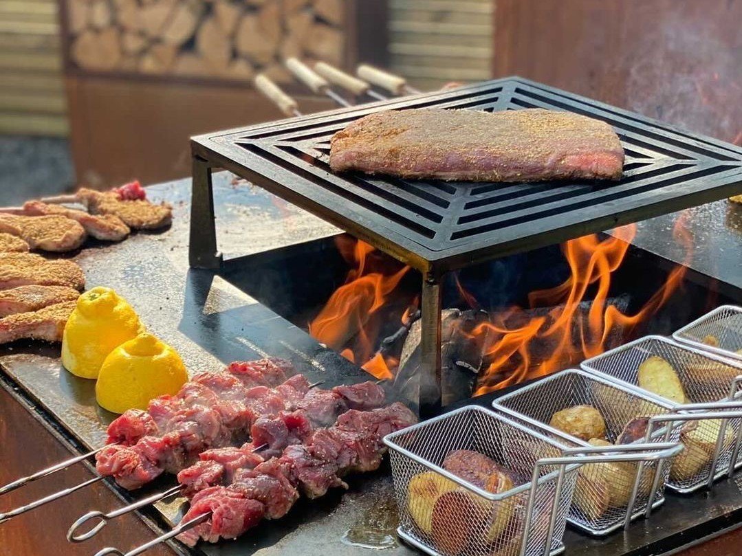 byJEMA Feuerstelle aus Cortenstahl mit sichtbaren Flammen – Fleisch, Grillspieße und Kartoffeln über offenem Feuer