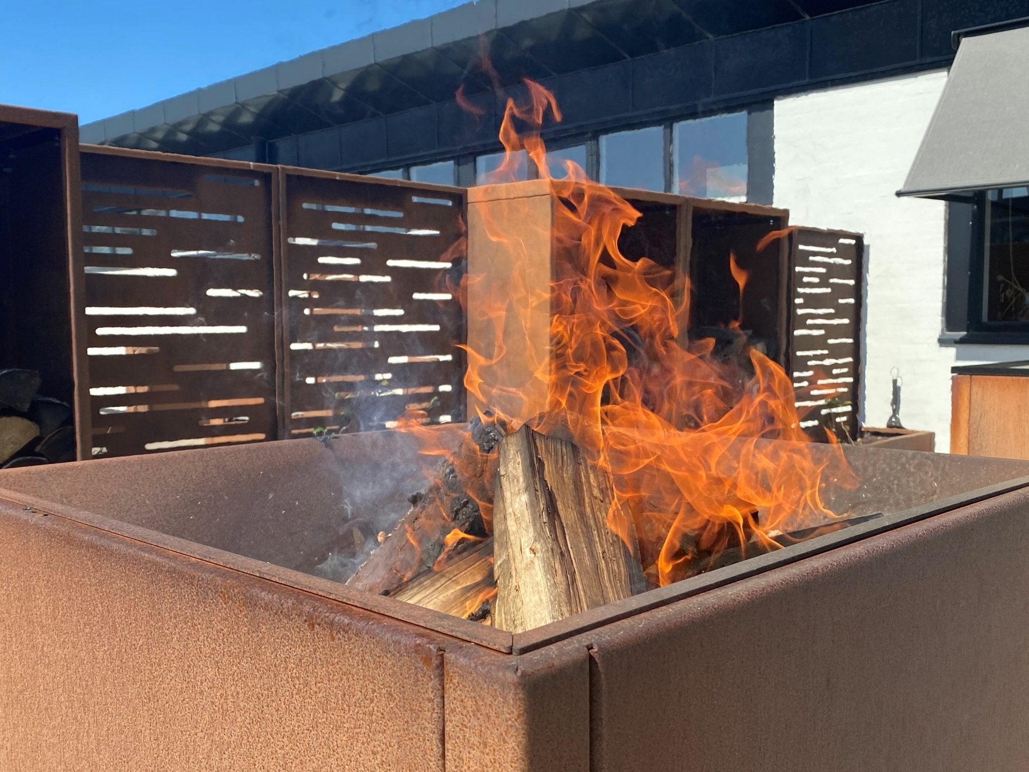Quadratische Feuerschale aus Cortenstahl mit brennendem Holzfeuer auf moderner Terrasse
