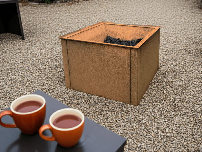 Quadratische Feuerschale aus Cortenstahl ohne Feuer auf Kiesflaeche im Garten