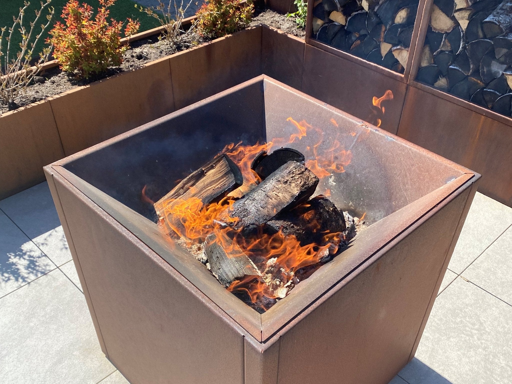 Quadratische Feuerstelle aus Cortenstahl mit brennendem Holz auf moderner Terrasse – Detailaufnahme