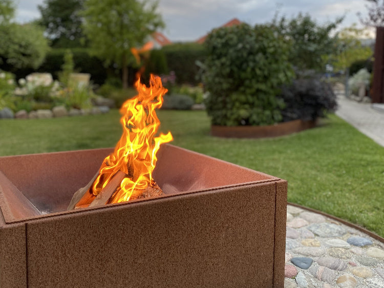 Quadratische Feuerschale aus Cortenstahl mit lebendigem Feuer, platziert in einem gepflegten Garten mit Rasen und Pflanzbeeten.