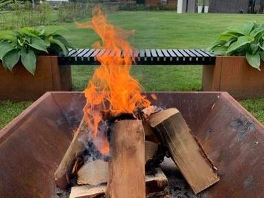 Quadratische Feuerstelle aus Cortenstahl mit brennendem Holz und hohen Flammen, aufgestellt im Garten mit Sitzbank und Pflanzkästen im Hintergrund.