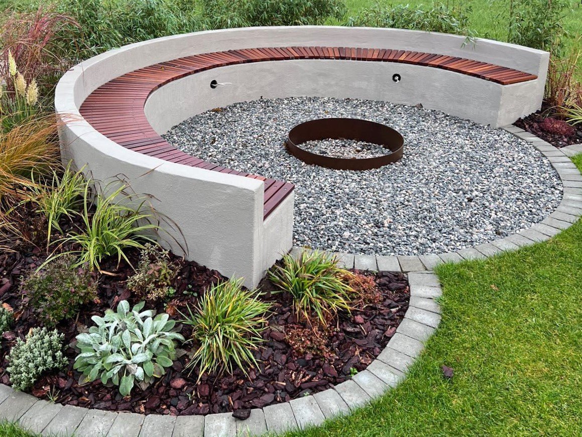 Runde Sitzanlage im Garten mit integrierter Holzsitzfläche und runder Feuerring aus Cortenstahl, umgeben von Kies, Stauden und Rasen.