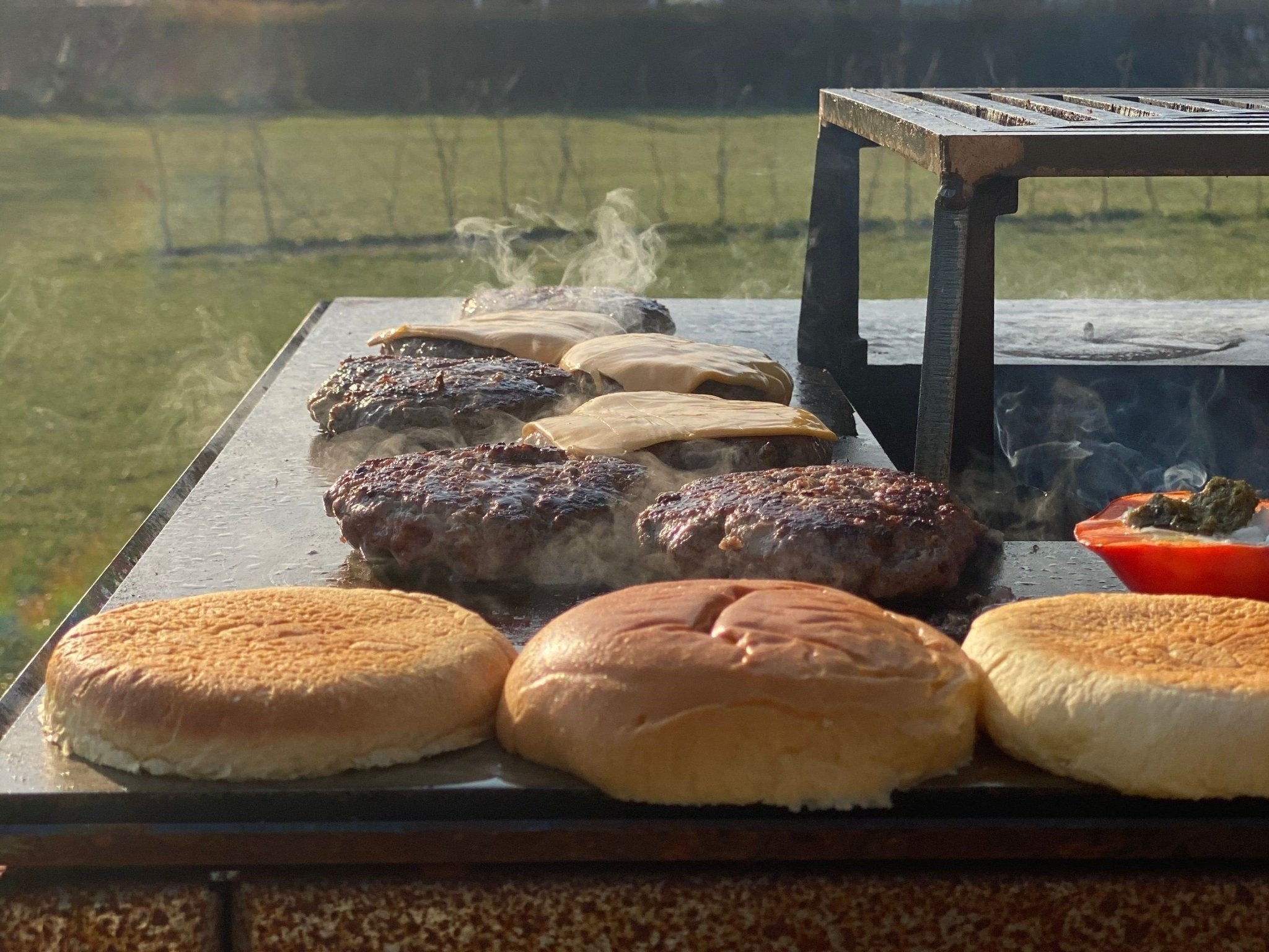 byJEMA Feuerplatte aus Cortenstahl mit Grillaufsatz – saftige Burger-Patties mit geschmolzenem Käse und Brioche-Brötchen beim Grillen im Garten