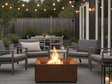 Rechteckige Feuerstelle aus Cortenstahl auf moderner Holzterrasse, umgeben von Loungemöbeln und Lichterkette im Garten
