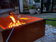 Quadratische Feuerschale aus Cortenstahl mit offenem Feuer, an dem Marshmallows auf Spießen geröstet werden – stimmungsvolle Gartenszene am Abend.