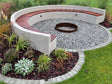 Runde Sitzanlage im Garten mit integrierter Holzsitzfläche und runder Feuerschale aus Cortenstahl, umgeben von Kies, Stauden und Rasen.